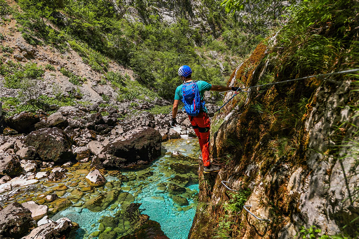 A climber in a blue helmet clipped to the steel cable above the turquoise Piva.